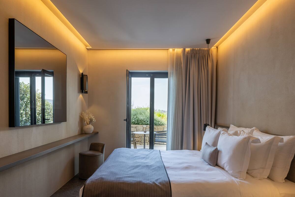 Ein modernes Hotelzimmer mit einem großen Bett, einem Spiegel, einem kleinen Tisch mit einer Vase und Blick auf Grünanlagen durch eine Schiebetür.