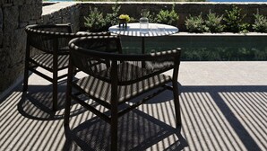 A covered outdoor seating area with two wicker chairs and a table, overlooking a scenic view of the sea and hills. - Noima Boutique Hotel Mykonos (Mykonos)