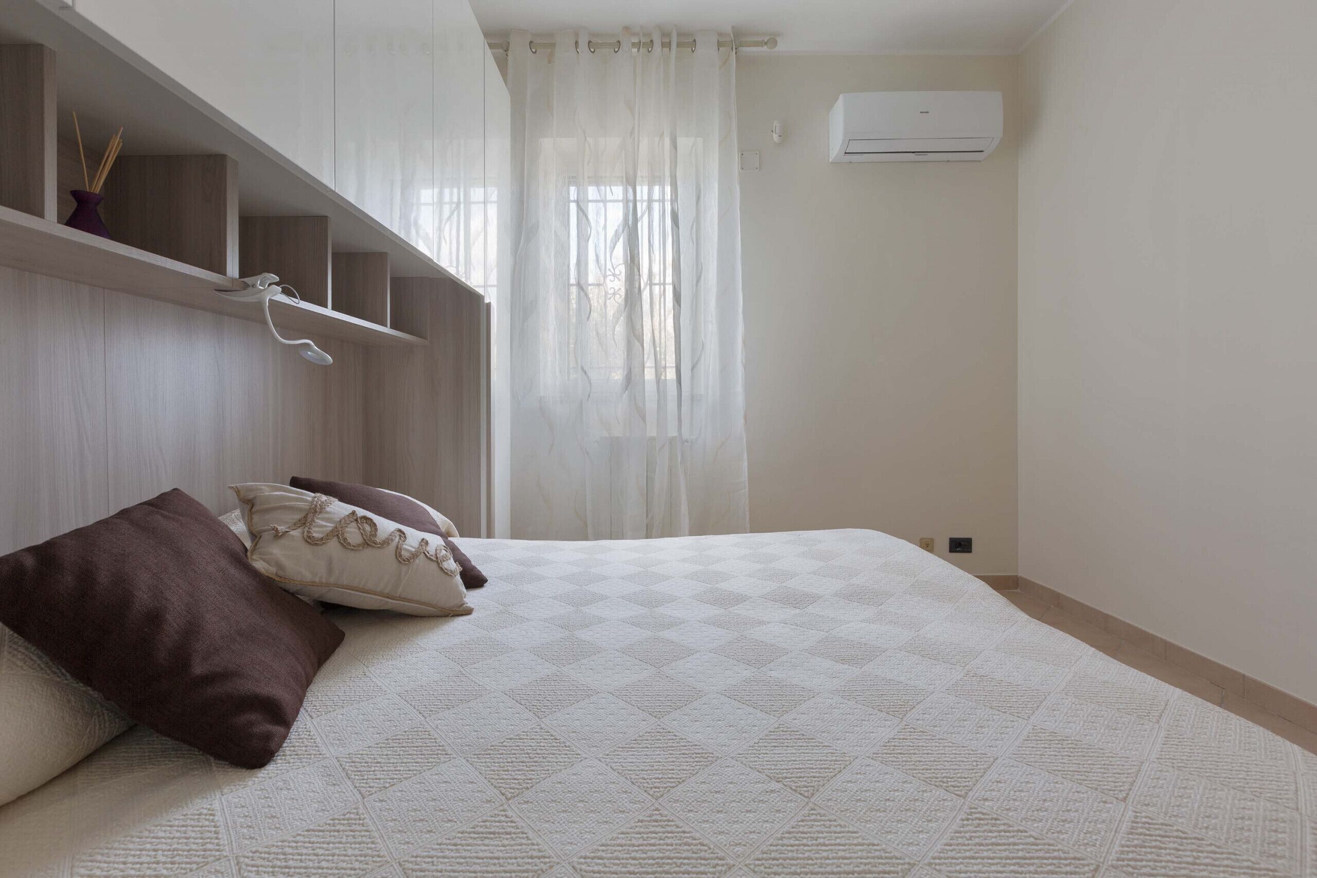 A bedroom with a bed, a window with sheer curtains, a wall-mounted air conditioner, and a shelf with decorative items.