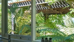 Et køkken med et stort vindue der udover en frodig have med bregner og en træpergola.