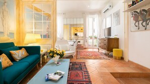Ein Wohnzimmer mit einer Couch, Couchtisch und Fernseher. Es gibt einen Essbereich mit Tisch und Stühlen. Das Zimmer verfügt über ein großes Fenster und ein Bild an der Wand.