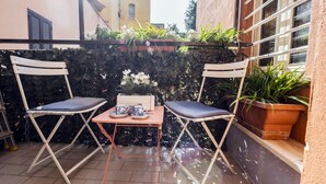 Uma pequena área externa com assentos para dois, incluindo cadeiras dobráveis e uma mesa, cercada por plantas e um muro de pedra.