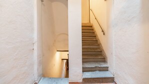 Un couloir étroit avec un escalier menant à l’étage supérieur. Les murs sont peints en blanc et une porte en bois se trouve au fond du couloir.