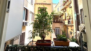 Une ruelle étroite en milieu urbain, bordée de bâtiments des deux côtés, avec des balcons ornés de plantes et de mobilier d’extérieur.