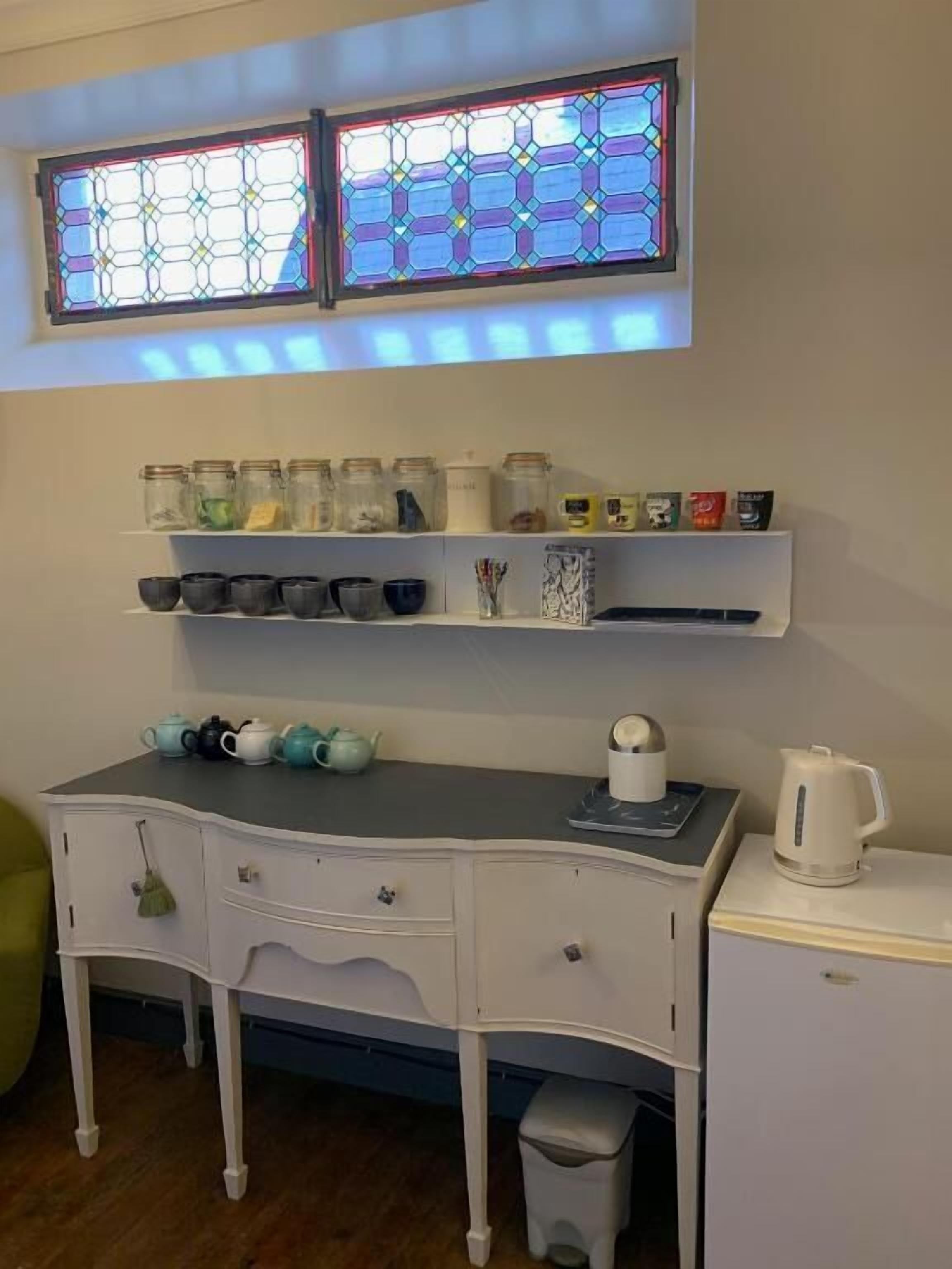 A kitchen with a white countertop, two shelves above containing jars and kitchenware, and a window with stained glass above.