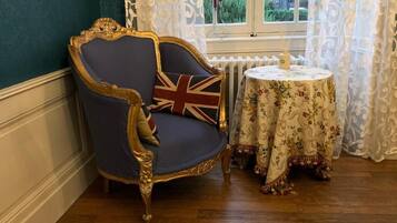 Een kamer met een donkerblauwe fauteuil met een Union Jack kussen, een ronde tafel met een tafelkleed met bloemenprint, en een raam met witte gordijnen.