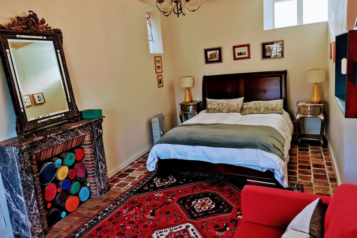 A hotel room with a bed, a large mirror, a chandelier, a red sofa, and a patterned rug.