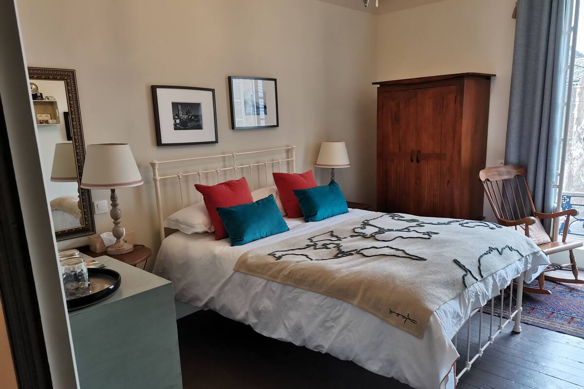 A neatly arranged bedroom with a bed, bedside lamps, a wooden wardrobe, and a chair by the window.
