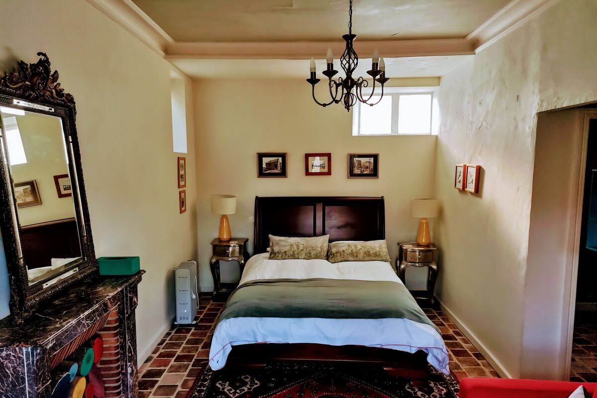 A hotel room with a large bed, two bedside lamps, a chandelier, a mirror, and a red chair.