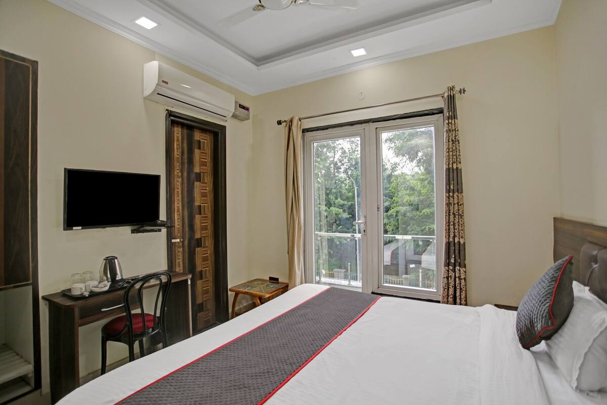 A hotel room with a bed, a desk with a chair, a television, an air conditioning unit, and a window with curtains.