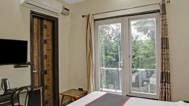 A hotel room with a bed, a desk with a chair, a television, an air conditioning unit, and a sliding glass door with a view of greenery outside. - Imperial Hotel Noida (Noida)