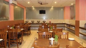 A restaurant with multiple wooden tables and chairs, a ceiling fan, and a television mounted on the wall. - Imperial Hotel Noida (Noida)