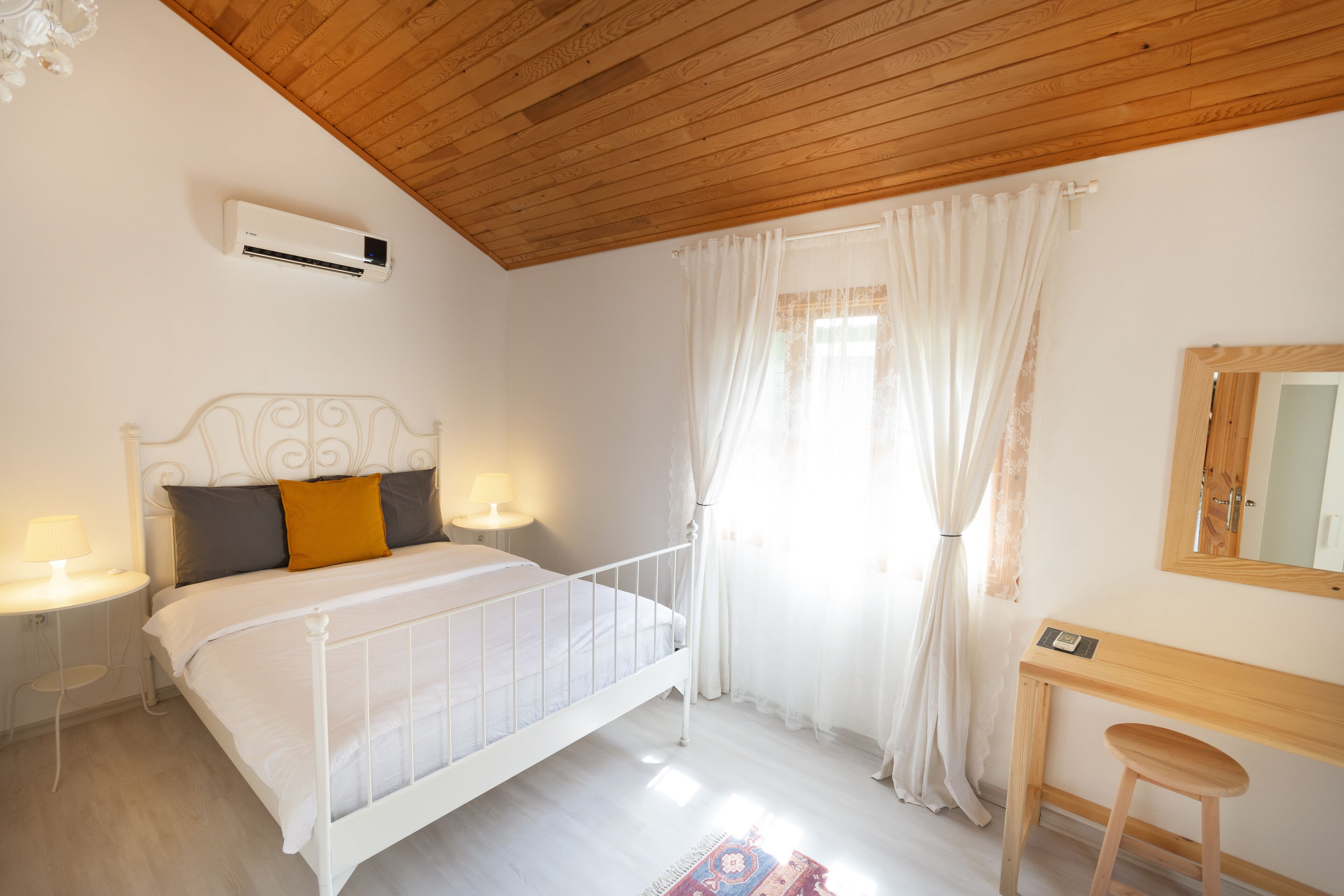 A bedroom with a white bed, a wooden desk, a mirror, and a window with white curtains.