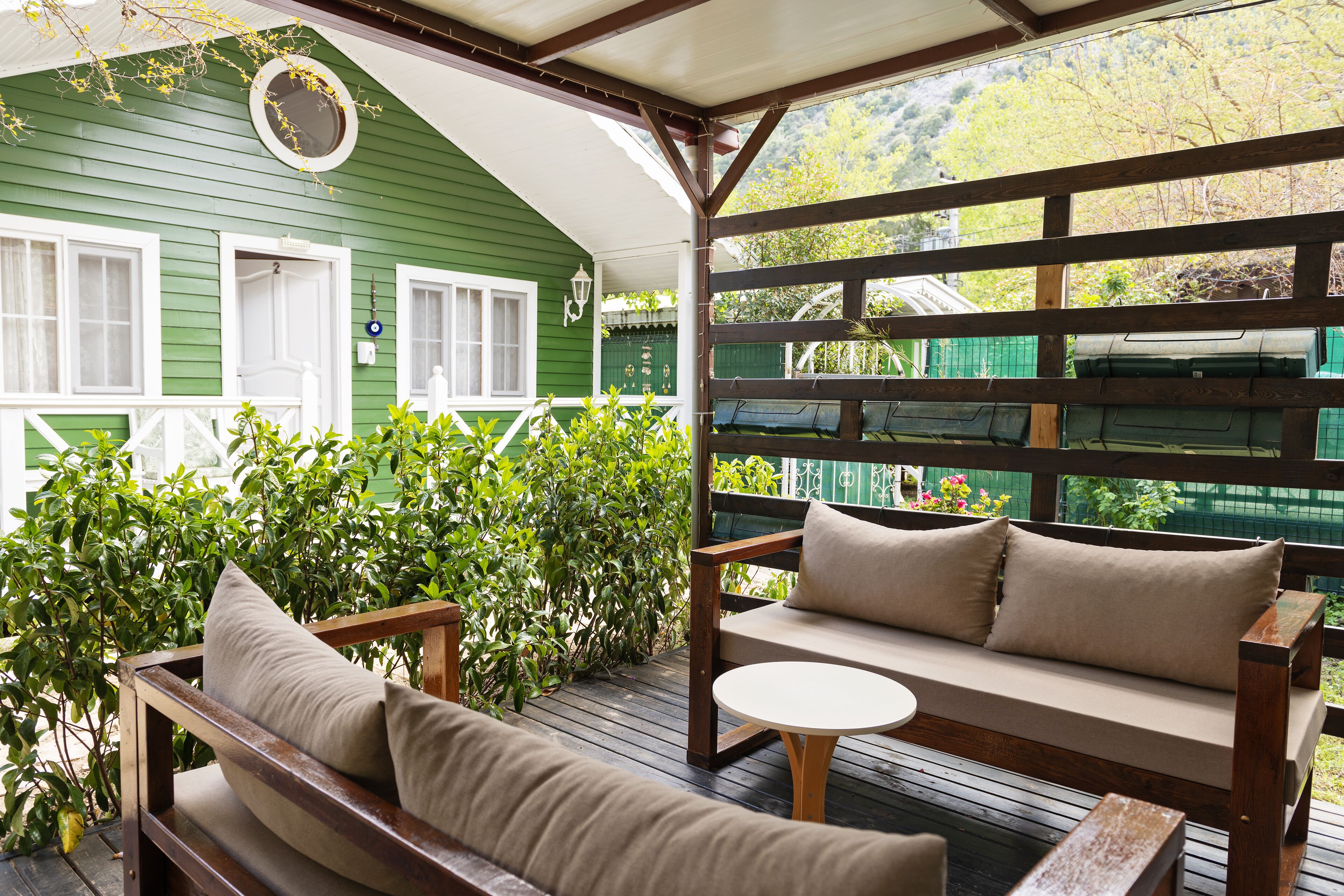 Una terraza de madera con una casa verde al fondo, un banco de madera y una mesa redonda.
