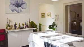 A hotel room with a bed, a desk with plants and a wine bottle, a large flower painting, and a door leading to a bathroom. - CASA GRANDE ALCALA HISTORIC CENTER (Catania)