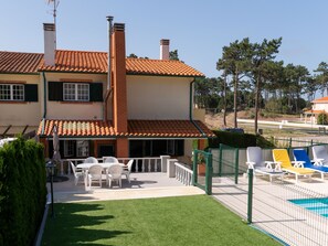Ein zweistöckiges Haus mit Ziegeldach, einem Balkon mit weißen Stühlen und einem eingezäunten Poolbereich.