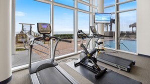 Un gimnasio con cintas de correr, bicicletas estáticas y una máquina de remo, con vistas a una piscina y zona exterior.