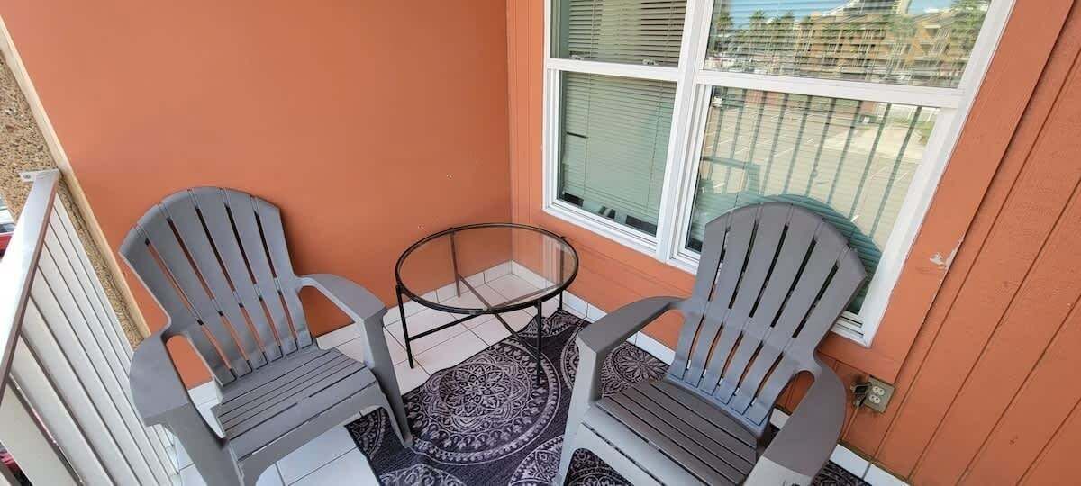 A small outdoor seating area with two Adirondack chairs and a glass table, adjacent to a window with a view of the neighborhood.