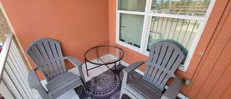 A small outdoor seating area with two Adirondack chairs and a glass table, adjacent to a window with a view of the neighborhood.