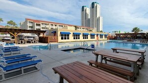 A pool area with lounge chairs and wooden tables, a building with a red roof, and a tall structure in the background. - Private Beach Access Pool & Hot Tub Bbq Pits 1 Bedroom Home by RedAwning (South Padre Island)