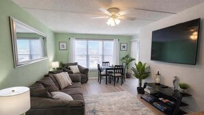 A living room with a sofa, a dining table, chairs, a ceiling fan, and a TV mounted on the wall. - Private Beach Access Pool & Hot Tub Bbq Pits 1 Bedroom Home by RedAwning (South Padre Island)