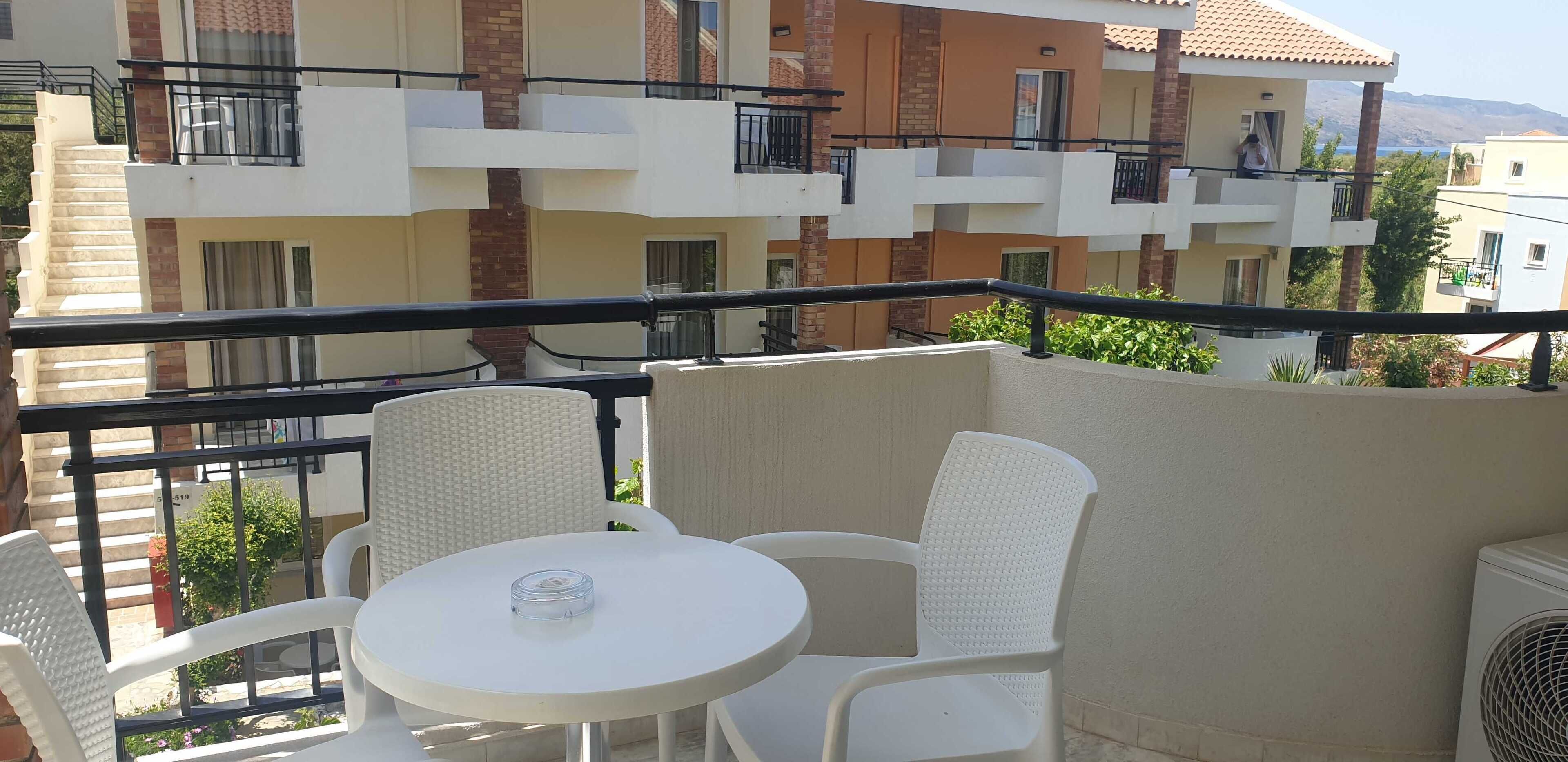 Un balcón con sillas de mimbre blanco y una pequeña mesa redonda, con vistas a una zona residencial con edificios de varios pisos y una perspectiva despejada del paisaje circundante.