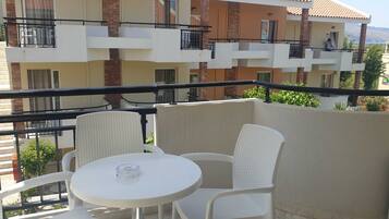 Un balcón con sillas de mimbre blanco y una pequeña mesa redonda, con vistas a una zona residencial con edificios de varios pisos y una perspectiva despejada del paisaje circundante.