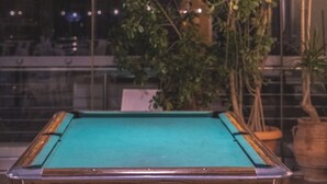 Une table de billard avec un cadre en bois et un drap de jeu vert, entourée de plantes en pot et d’un luminaire suspendu.