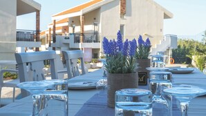 Un espace extérieur aménagé pour prendre les repas, avec une table dressée pour deux personnes, un pot de fleurs aux fleurs bleues et, en arrière-plan, un bâtiment avec un toit recouvert de tuiles.
