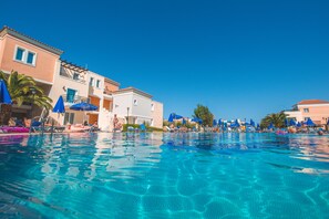 Una piscina con agua cristalina y azul, rodeada de sillas de descanso y sombrillas, con edificios colindantes con balcones.