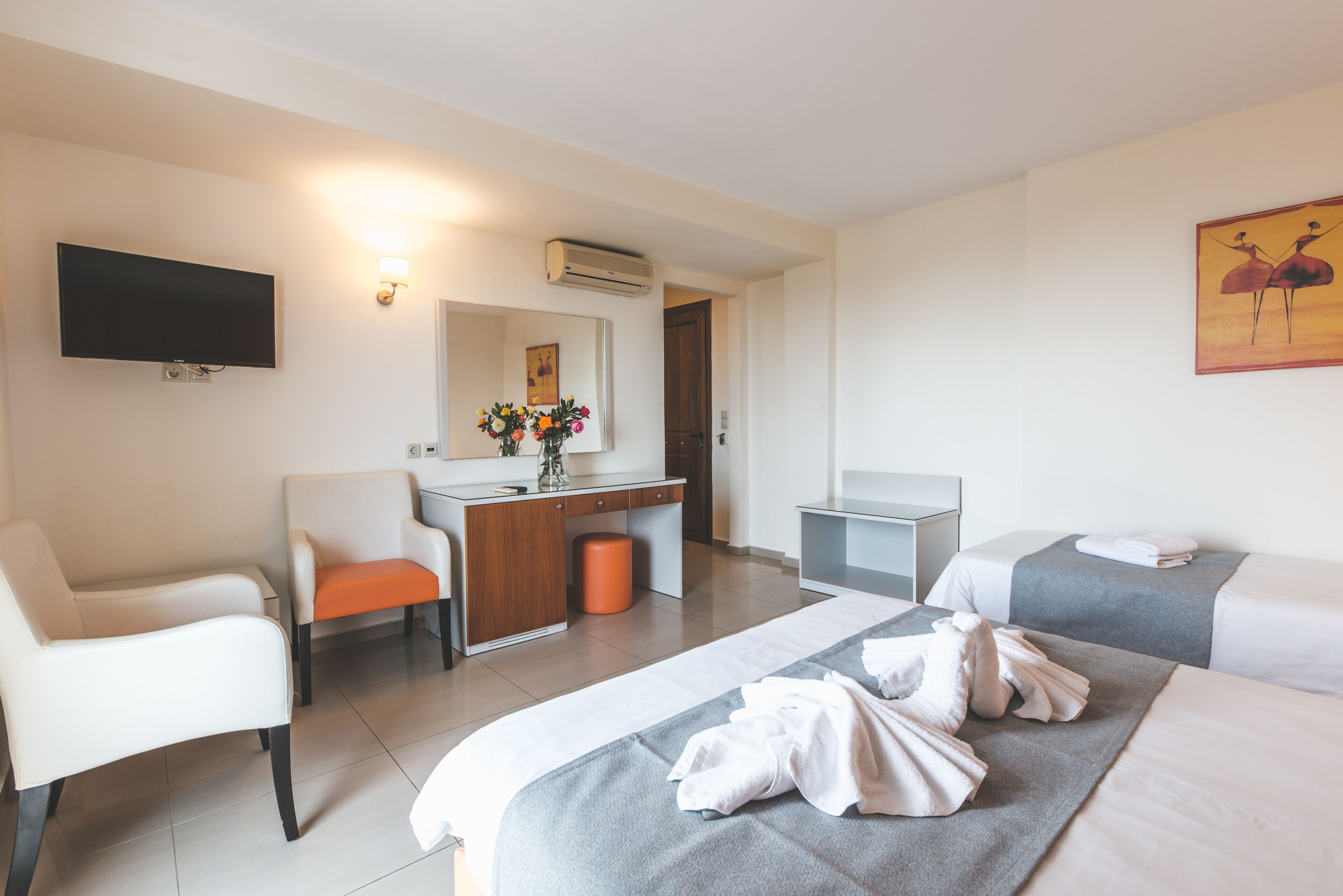 Habitación de hotel con una cama, dos sillas blancas, un escritorio con una computadora, un televisor y un cuadro en la pared.