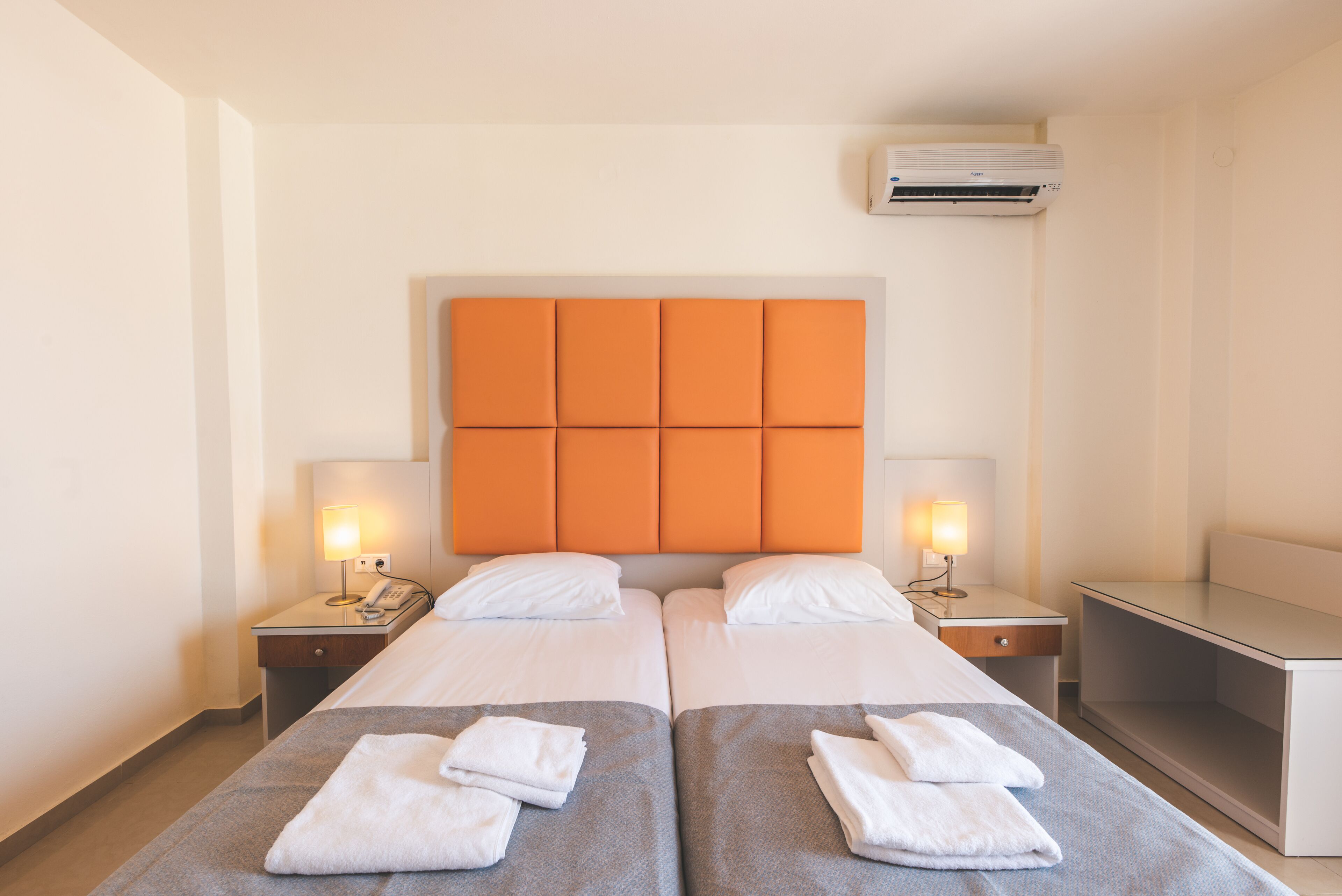 A hotel room with two beds, a headboard, bedside tables with lamps, and an air conditioning unit.