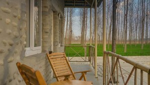 A covered outdoor seating area with wooden chairs and tables, a stone wall, and a view of a green field and trees. - Anand at the Satluj (Anandpur Sahib)