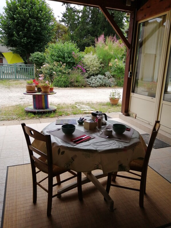 A round table with chairs, a bowl, a teapot, and a cup on a covered patio with a garden view.