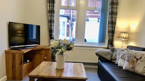 Un salon avec un téléviseur posé sur un meuble en bois, une table basse ornée d’un vase de fleurs, un canapé avec des coussins à motifs et une fenêtre avec des rideaux.