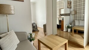 Un salon moderne avec un canapé, une table en bois et un miroir qui reflète la pièce. On y trouve une lampe, une petite plante et un tableau accroché au mur.