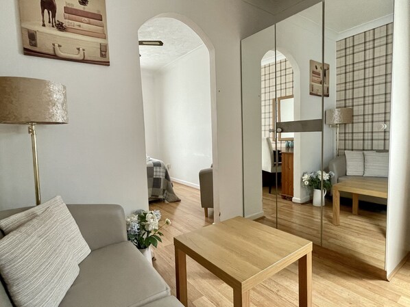 A modern living room with a sofa, a wooden table, and a mirror reflecting the room. There is a lamp, a small plant, and a painting on the wall.