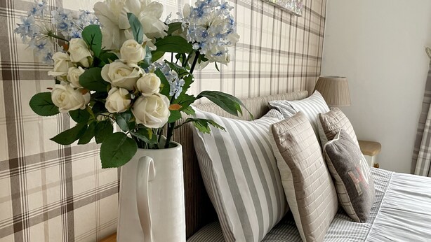 Une chambre à coucher avec une tête de lit à motifs écossais, une table de chevet en bois, un vase blanc contenant des fleurs blanches et bleues, et une literie à rayures.