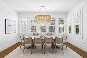 A dining room with a large table, six chairs, a chandelier, and large windows with blinds.