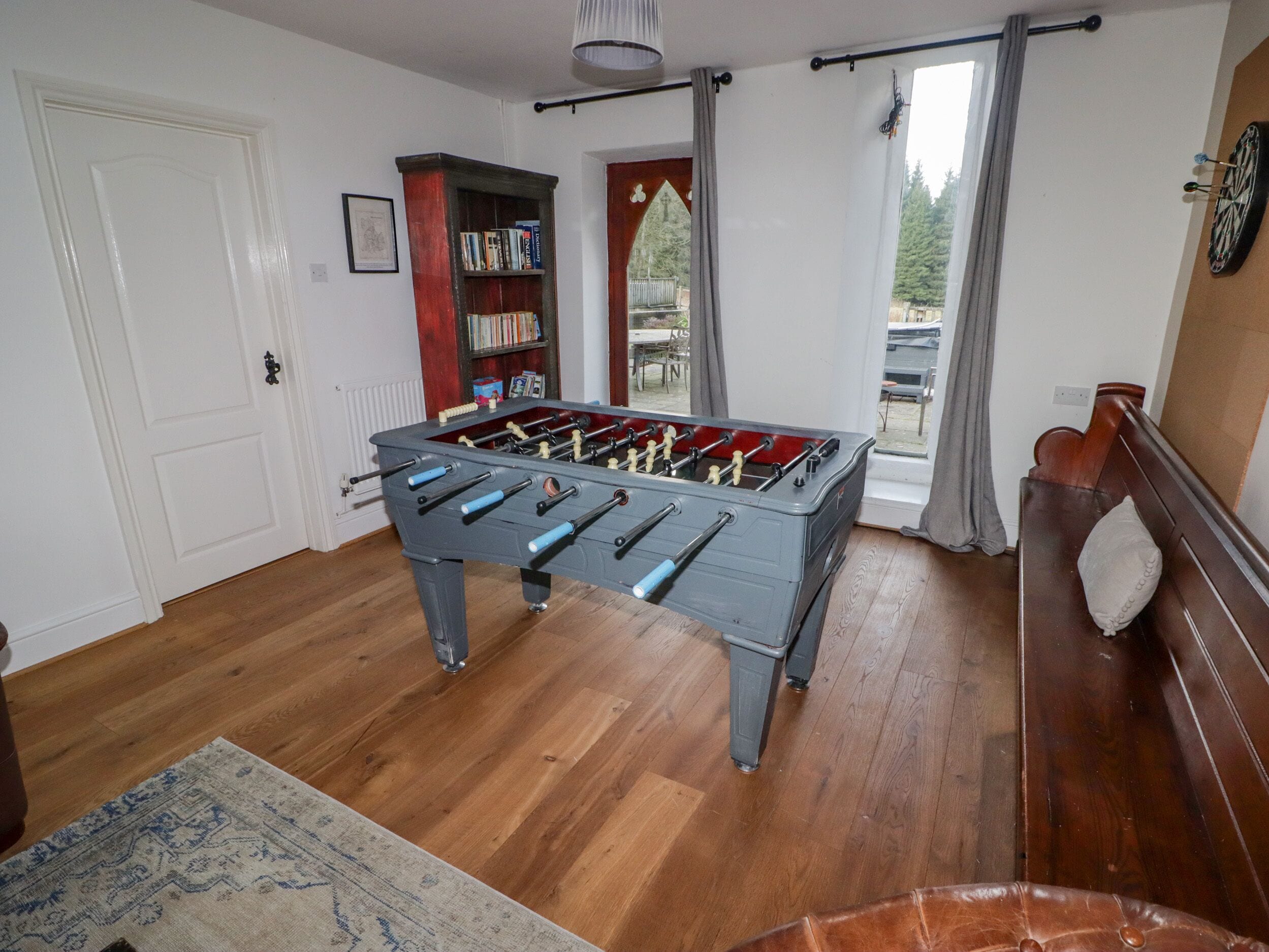 Una habitación con mesa de fútbol de mesa, piso de madera, una estantería y un ventanal con cortinas.