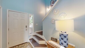 Un couloir avec une porte blanche, un escalier en bois, un mur blanc orné d’assiettes décoratives et une lampe à motifs posée sur une table en bois.
