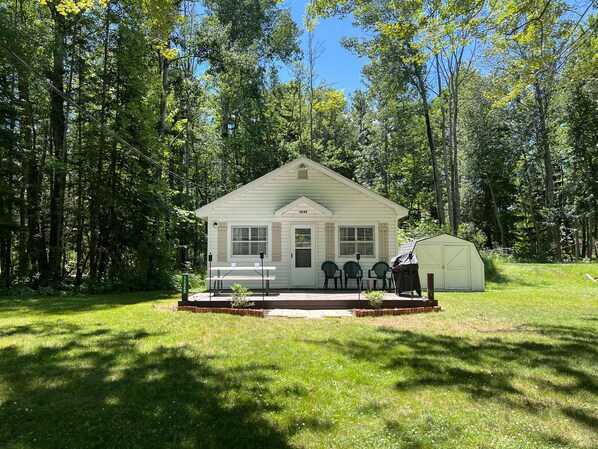 A small, single-story cabin with a porch and a covered area, surrounded by a forest.