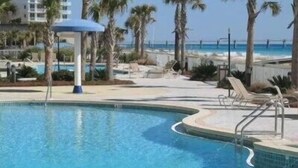 A swimming pool with palm trees and lounge chairs, surrounded by a building with a view of the ocean. - Azure 222 — Renovated 3BR Gulf-Side Condo with Beach Service (Fort Walton Beach)