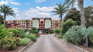 A hotel entrance with a brick pathway, palm trees, and a building with a central archway. - Azure 222 — Renovated 3BR Gulf-Side Condo with Beach Service (Fort Walton Beach)