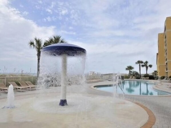 Ein Trinkbrunnen mit kreisförmigem Becken, einem Pool und Palmen im Hintergrund.