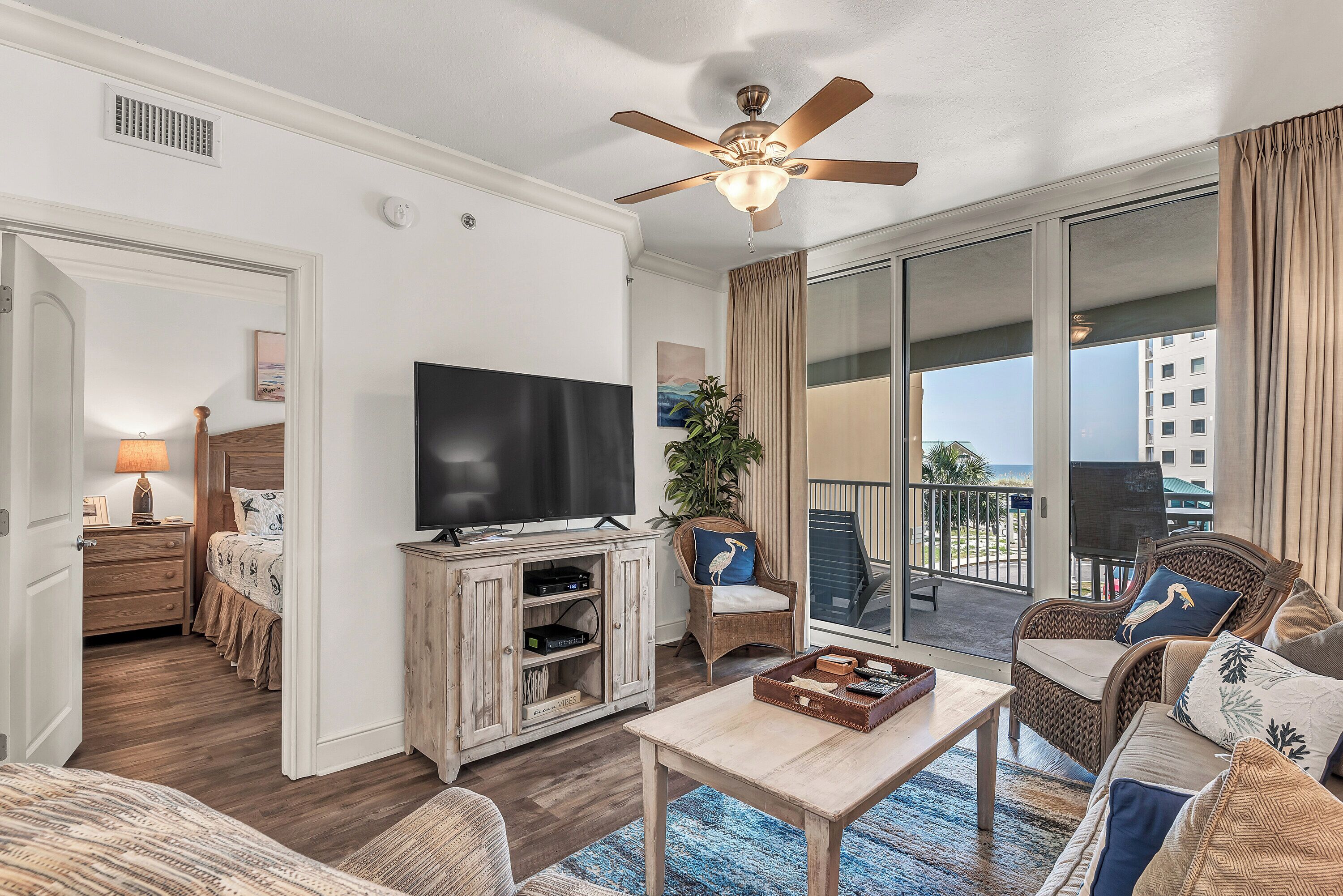 Ein Wohnzimmer mit Deckenventilator, Fernseher, Holzschrank, Sofa, Sessel und einem Balkon mit Blick.