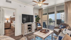 A living room with a ceiling fan, a TV, a wooden cabinet, a sofa, a chair, and a balcony with a view. - Azure 222 — Renovated 3BR Gulf-Side Condo with Beach Service (Fort Walton Beach)