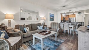 A living room with a sofa, armchairs, a coffee table, and a dining area with chairs and a table. There is a kitchen in the background with appliances and a clock on the wall. - Azure 222 — Renovated 3BR Gulf-Side Condo with Beach Service (Fort Walton Beach)
