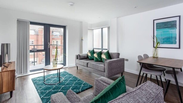 Una sala moderna con un sofá gris, una mesa de centro y una mesa de comedor con sillas. Hay un ventanal con vistas a edificios.