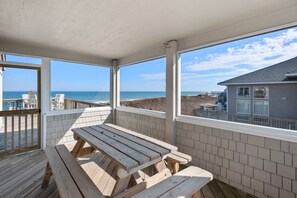 Un espacio al aire libre techado con una mesa de picnic de madera y bancos, amplias ventanas que dan a vistas de la playa y el océano.